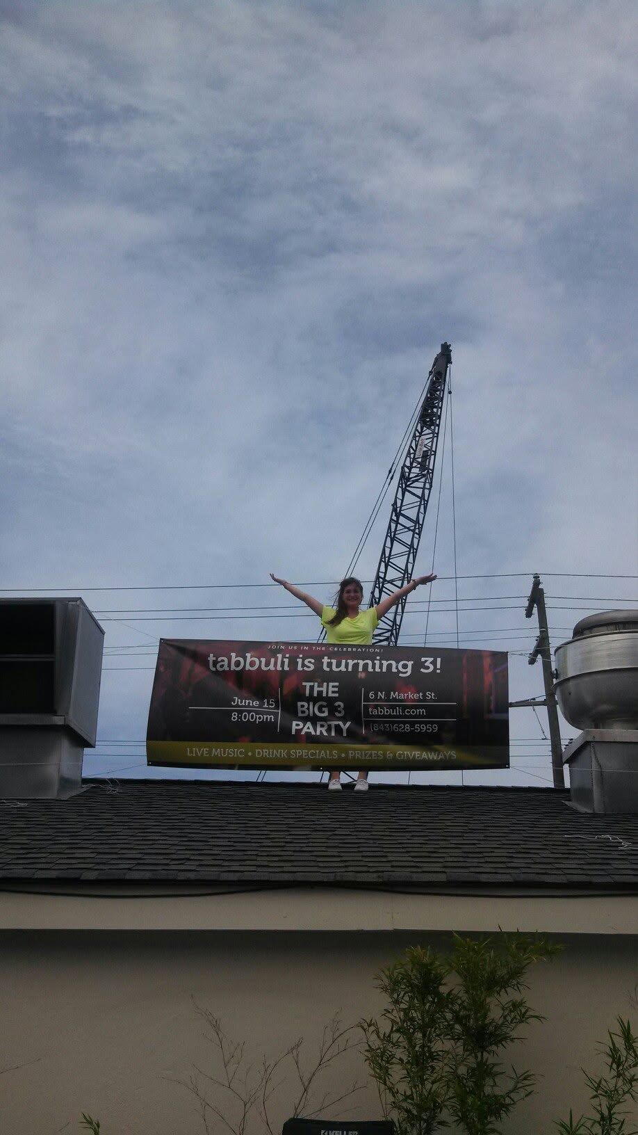 Brianna on the rooftop of Tabbuli in Charleston, SC, hanging a 'Tabbuli is turning 3!' anniversary banner.