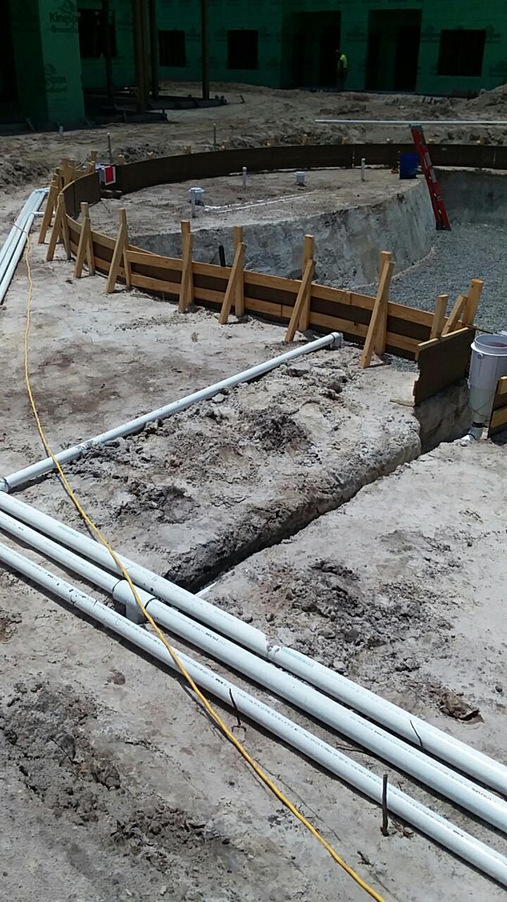 Pool shell shotcrete and plumbing layout progress with surrounding building structure in the background.
